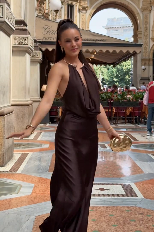 Woman in a brown dress standing in an elegant indoor setting with decorative columns and chandeliers.