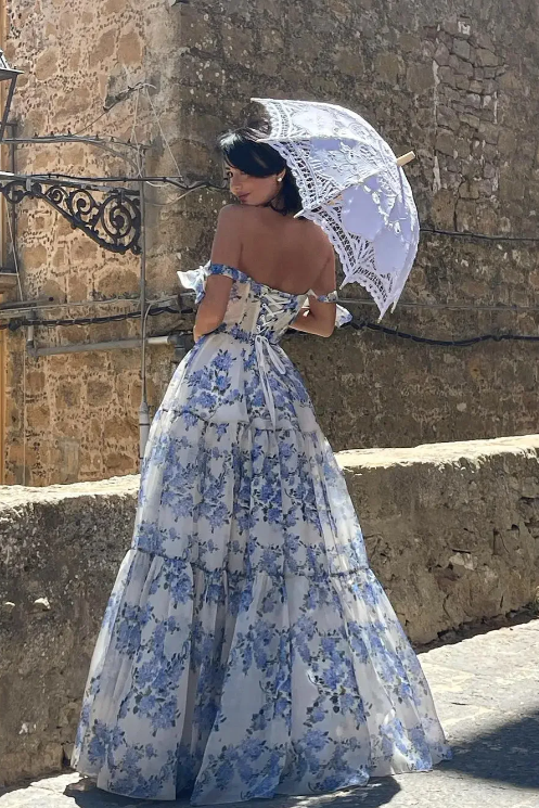 Woman in a blue floral dress holding a white lace umbrella against a stone wall.