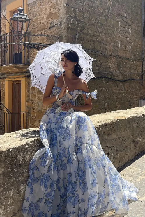 Woman in a blue floral dress holding a white parasol in an old stone street.