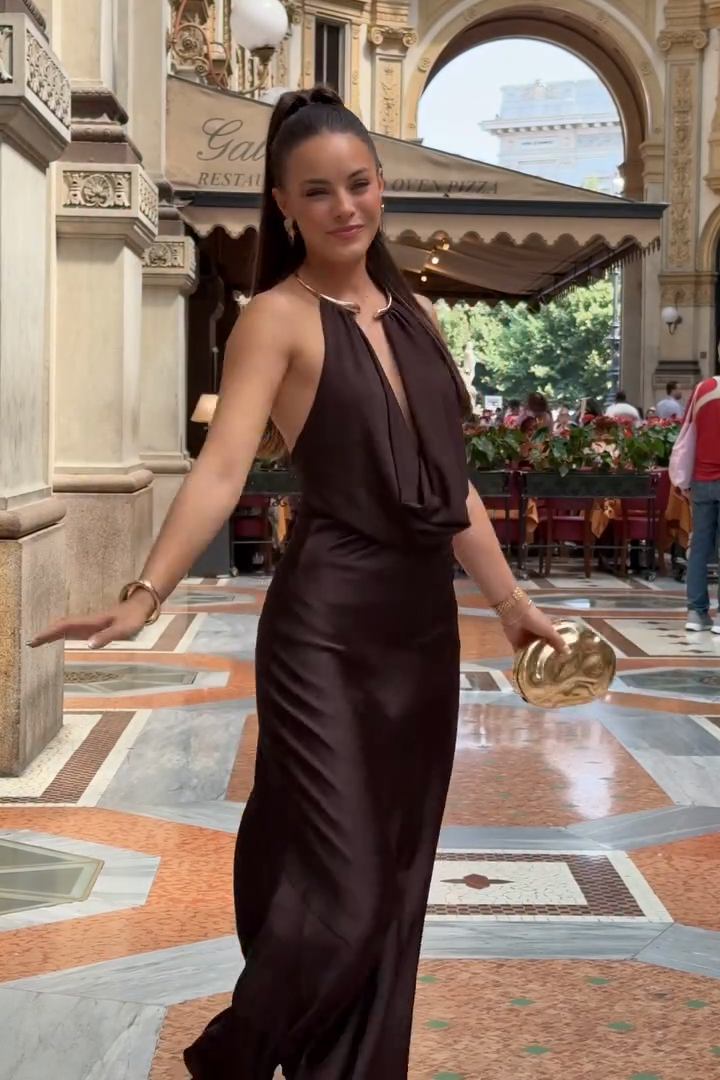 Woman in a brown dress standing in an elegant indoor setting with decorative columns and chandeliers.