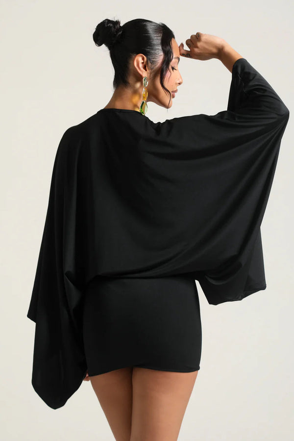 Woman wearing a black outfit with a cape-like garment on a plain background